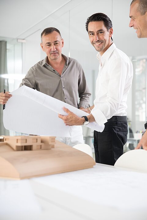 Drei Architekten begutachten Baupläne in einem Büro, auf dessen Tisch ein Holzmodell steht.
